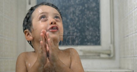 Téléchargez les photos : Joyeux enfant profitant de l'heure du bain avec des gouttelettes tombant en super 800 ips au ralenti, jeune garçon rayonnant de joie en observant le débit d'eau debout dans la douche - en image libre de droit