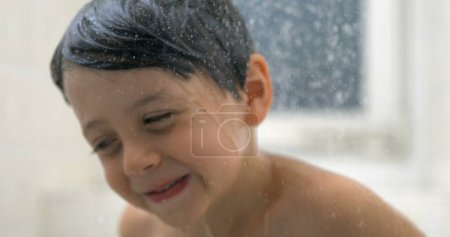 Téléchargez les photos : Joyeux visage d'enfant en gros plan pendant le bain avec des gouttelettes d'eau qui coulent au ralenti dans la douche - en image libre de droit