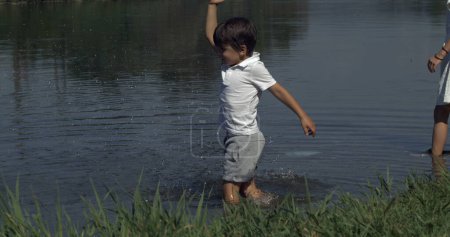 Foto de Niño pequeño salpicando agua con la mano por el lago capturado. Niño disfrutando de la naturaleza conectada con el mundo natural - Imagen libre de derechos
