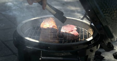 Téléchargez les photos : Mettre un morceau de viande dans un barbecue portable avec spatule pendant l'activité de camping Wekend capturé fumée blanche - en image libre de droit