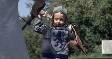 Téléchargez les photos : Jeune chevalier en tenue médiévale balançant l'épée avec bouclier, enfant en costume d'époque s'engageant dans un jeu d'épée capturé à grande vitesse avec rampe de vitesse - en image libre de droit