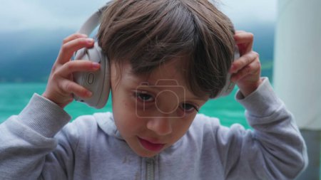 Photo for One cute little boy putting on headphones overlooking lake in background. Portrait of happy child wearing over the ear headphone, 4 year old kid - Royalty Free Image