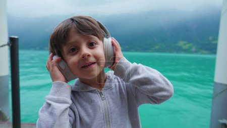 Téléchargez les photos : Adorable petit garçon écoutant de la musique avec lac pittoresque en arrière-plan. Enfant de 4 ans rebondissant au rythme de la chanson, mains tenant un casque - en image libre de droit