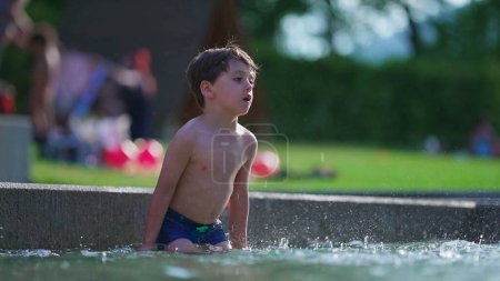 Téléchargez les photos : Joyeux enfant éclaboussant l'eau à la piscine extérieure au ralenti. Petit garçon jetant de l'eau dans les airs avec la main dans 120fps pendant la journée d'été - en image libre de droit