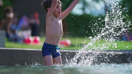 Téléchargez les photos : Joyeux enfant éclaboussant l'eau à la piscine extérieure au ralenti. Petit garçon jetant de l'eau dans les airs avec la main dans 120fps pendant la journée d'été - en image libre de droit