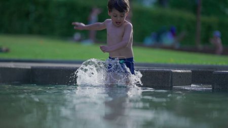 Téléchargez les photos : Enfant heureux courant dans l'eau de la piscine au ralenti pendant la journée d'été. Petit garçon actif éclaboussant l'eau coule vers l'avant en 120fps - en image libre de droit