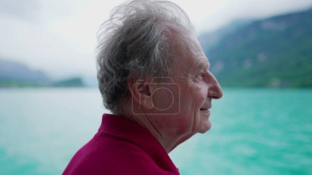 Téléchargez les photos : Homme âgé insouciant profitant de la vue sereine de la nature sur le lac et les montagnes tout en voyageant en bateau, profil en gros plan de la personne âgée dans les années 70 contempler et explorer de nouveaux horizons - en image libre de droit