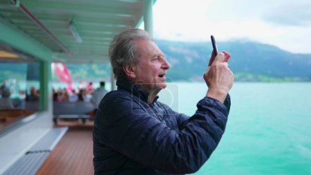 Téléchargez les photos : Homme âgé voyageant en bateau tirant sur le téléphone portable pour prendre une photo du lac et vue sur la montagne. Homme plus âgé homme retraité explorer de nouveaux horizons filmer la nature avec un appareil smartphone - en image libre de droit