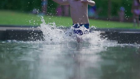 Foto de Niño feliz corriendo en el agua de la piscina en cámara lenta durante el día de verano. Niño activo salpicando agua corre hacia adelante en 120fps - Imagen libre de derechos