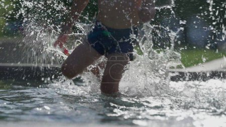 Foto de Niño divirtiéndose en la piscina pública durante el caluroso día de verano con gotas de agua salpicando en cámara lenta, niño saliendo junto a la piscina - Imagen libre de derechos