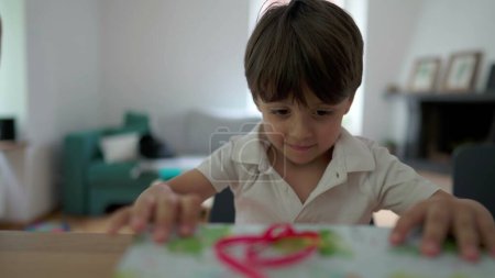 Foto de Emocionado niño abriendo presente en casa. Un niño pequeño desenvolviendo regalo, papel desgarrador, ansiosa anticipación de premio - Imagen libre de derechos