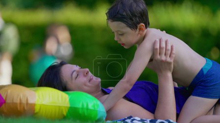 Téléchargez les photos : Mère heureuse et l'enfant couché dans l'herbe extérieure après avoir nagé à la piscine. Petit garçon et maman attachement affectueux et embrasser moment - en image libre de droit