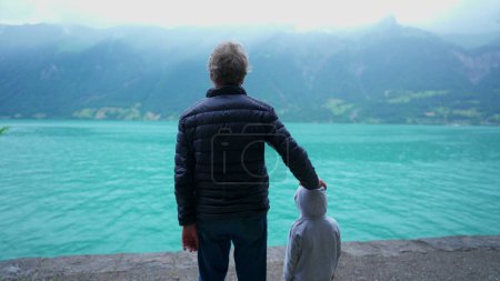 Téléchargez les photos : Dos de grand-père et petit-fils debout devant le lac et vue sur les montagnes. Famille profitant du grand air portant des vestes sous la pluie bruine, Scène calme et paisible - en image libre de droit