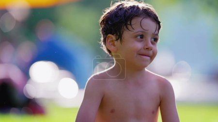 Téléchargez les photos : Joyeux jeune garçon assis dehors après avoir nagé à la piscine souriant. Enfant torse nu profitant d'une journée chaude d'été - en image libre de droit