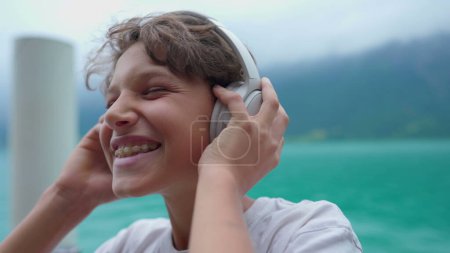 Foto de Joven alegre escuchando música usando auriculares junto al lago. Cara de primer plano de un adolescente sosteniendo auriculares sobre la oreja rebotando al ritmo de la canción - Imagen libre de derechos