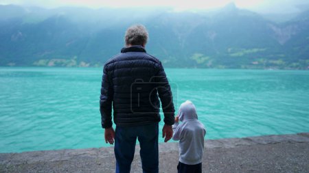 Foto de Parte posterior del abuelo y el nieto de pie frente a la vista del lago y las montañas. Familia disfrutando del aire libre con chaquetas en la lluvia llovizna, calma y escena pacífica - Imagen libre de derechos