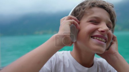 Photo for Joyful young boy listening to music wearing headphones standing by lake. Close-up face of teenager kid holding headphones over the ear bouncing head to the beat of song - Royalty Free Image