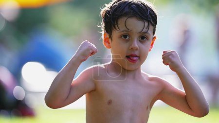 Téléchargez les photos : Un adorable petit garçon muscles fléchissants assis sur l'herbe à l'extérieur après avoir nagé à la piscine pendant la chaude journée d'été. Enfant montre de minuscules biceps - en image libre de droit