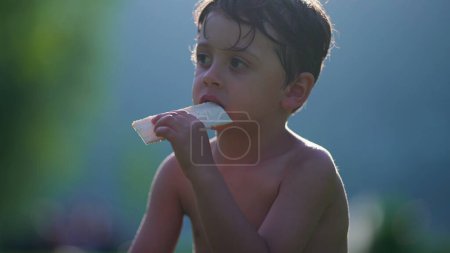 Photo for Pensive child eating biscuit while drying in the sun after playing at pool. Shirtless small boy enjoys summer vacations observing surroundings - Royalty Free Image