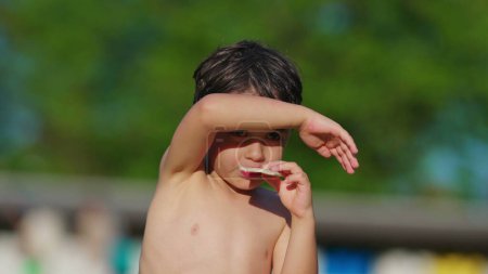 Téléchargez les photos : Enfant torse nu couvrant le visage et les yeux du soleil avec le bras tout en mangeant des biscuits, séchant au soleil après avoir nagé à la piscine pendant la chaude journée d'été - en image libre de droit