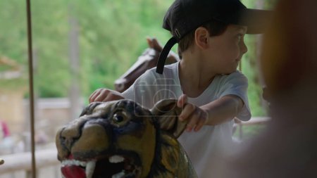Téléchargez les photos : Joyeux petit garçon chevauchant carrousel au parc, enfant profitant de la journée d'été assis sur le dessus du tigre dans le manège en mouvement - en image libre de droit