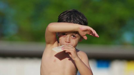 Foto de Niño sin camisa cubriendo la cara y los ojos del sol con el brazo mientras come galletas, secándose al sol después de nadar en la piscina durante el cálido día de verano - Imagen libre de derechos