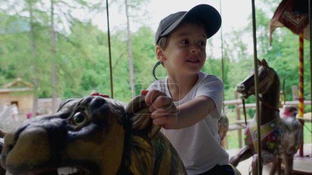 Téléchargez les photos : Petit garçon excité monte carrousel au parc, enfant s'amusant au manège en mouvement - en image libre de droit
