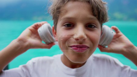 Foto de Retrato de un niño preadolescente feliz poniendo auriculares, cara de primer plano del niño adolescente escuchando audiolibro, podcast o canción con vistas al lago en el fondo - Imagen libre de derechos