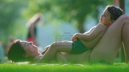 Téléchargez les photos : Mère et enfant couchés sur l'herbe au bord de la piscine pendant la journée d'été, moment affectueux de famille aimante franche, moment de style de vie de la maternité liant son fils - en image libre de droit