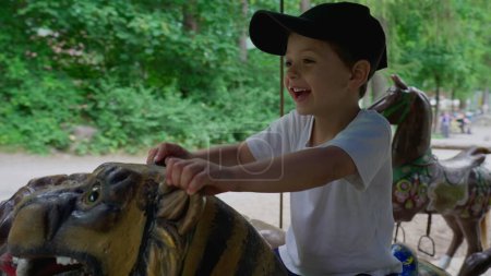 Téléchargez les photos : Joyeux petit garçon assis sur le carrousel dans le parc d'été - en image libre de droit