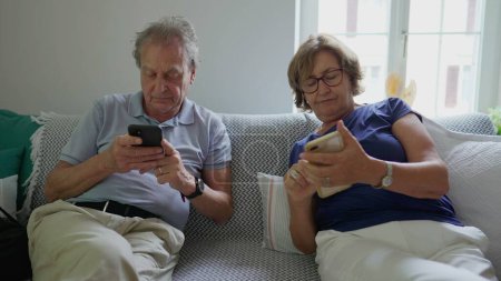 Photo for Senior married couple looking at their cellphones sitting at home couch. Elderly man and woman using modern technology, staring at screens, absorbed by smartphones - Royalty Free Image