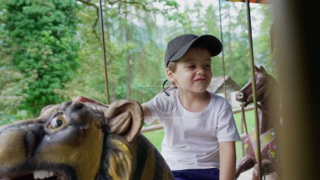 Téléchargez les photos : Petit garçon excité monte carrousel au parc, enfant s'amusant au manège en mouvement - en image libre de droit