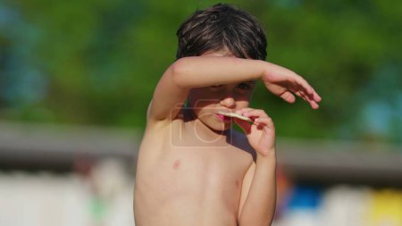 Foto de Niño sin camisa cubriendo la cara y los ojos del sol con el brazo mientras come galletas, secándose al sol después de nadar en la piscina durante el cálido día de verano - Imagen libre de derechos