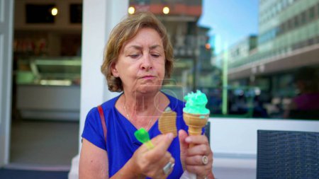 Téléchargez les photos : Senior Lady Relâcher un Froid-crème glacée traiter par une journée ensoleillée à Parlor Shop. Femme plus âgée savourant un dessert au cône de gaufre - en image libre de droit