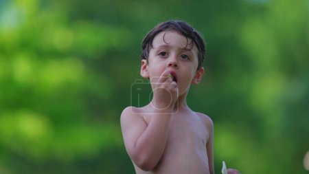 Téléchargez les photos : Un cher petit garçon debout à l'extérieur torse nu après avoir nagé à la piscine en mangeant des biscuits, enfant réfléchi avec un regard contemplatif séchant tout en prenant une bouchée de biscuit - en image libre de droit