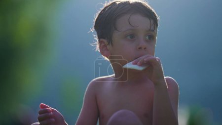 Téléchargez les photos : Enfant pensif manger des biscuits tout en séchant au soleil après avoir joué à la piscine. Petit garçon torse nu aime les vacances d'été en observant les environs - en image libre de droit