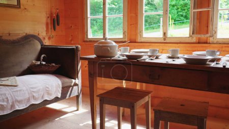 Foto de Entorno rústico de mesa de madera y ventana en chalet rural, sofá antiguo con vajilla blanca. Platos de cerámica humildes, tazas de té y utensilios en la mesa envejecida - Imagen libre de derechos