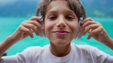 Foto de Retrato de un niño preadolescente feliz poniendo auriculares, cara de primer plano del niño adolescente escuchando audiolibro, podcast o canción con vistas al lago en el fondo - Imagen libre de derechos