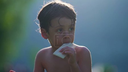 Téléchargez les photos : Enfant pensif manger des biscuits tout en séchant au soleil après avoir joué à la piscine. Petit garçon torse nu aime les vacances d'été en observant les environs - en image libre de droit