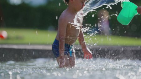 Téléchargez les photos : Petit garçon éclaboussant avec seau d'eau en 120 ips au ralenti à la piscine publique, activité amusante chaude de la journée d'été - en image libre de droit