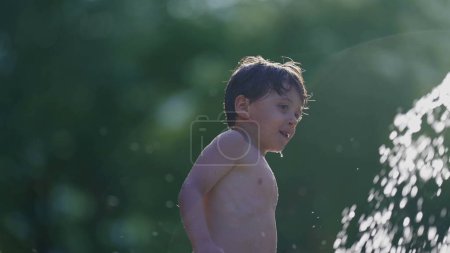 Téléchargez les photos : Petit garçon se rafraîchissant à la fontaine d'eau de la piscine publique au ralenti 120fps. Enfant profitant des éclaboussures de gouttelettes d'eau pendant la chaude journée d'été - en image libre de droit
