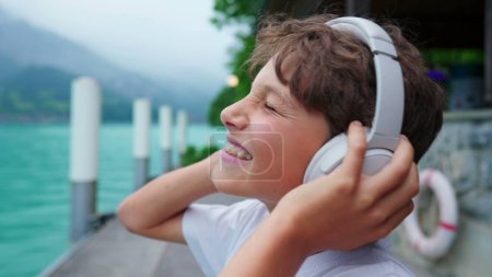 Foto de Joven adolescente absorto deslizándose en los auriculares, fondo junto al lago con niño absorbido en contenido de audio - Imagen libre de derechos