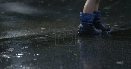Téléchargez les photos : Pieds d'enfant marchant sous la pluie à l'extérieur dans le trottoir de la rue de la ville avec des gouttelettes d'eau tombant au ralenti super capturé avec une caméra haute vitesse, jour de pluie - en image libre de droit