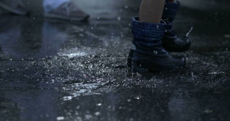Téléchargez les photos : Gros plan bottes de pluie marchant dans les flaques d'eau éclaboussant des gouttelettes partout à grande vitesse 800 ips pendant la soirée de l'après-midi pluvieux - en image libre de droit