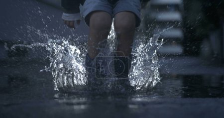 Foto de Un niño excitado con botas de lluvia salta al charco de agua en cámara lenta capturando gotas en el aire con una cámara de alta velocidad, temporada de lluvias - Imagen libre de derechos