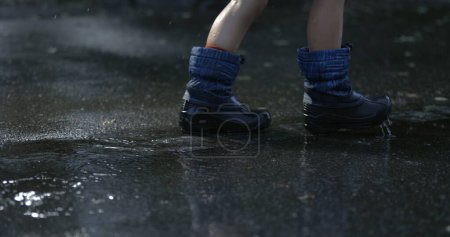 Téléchargez les photos : Pieds d'enfant marchant sous la pluie à l'extérieur dans le trottoir de la rue de la ville avec des gouttelettes d'eau tombant au ralenti super capturé avec une caméra haute vitesse, jour de pluie - en image libre de droit