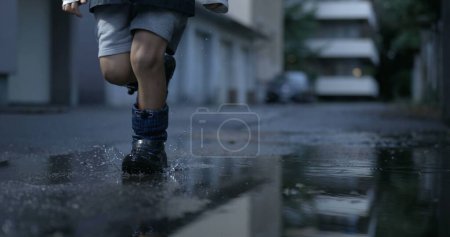 Téléchargez les photos : L'enfant court dans la flaque d'eau au ralenti. Gros plan gamin portant des bottes de pluie éclaboussant des gouttelettes après un après-midi pluvieux, scène d'enfance nostalgique - en image libre de droit