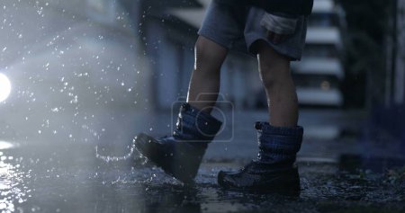 Téléchargez les photos : Enfant insouciant jouant avec la flaque d'eau dans les coups de pied de rue et les éclaboussures de gouttelettes dans l'air au ralenti super capturé avec une caméra à grande vitesse - en image libre de droit