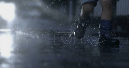 Foto de Niño corre en charco de agua con botas de lluvia durante la temporada de lluvias en cámara lenta con gotitas volando en el aire, concepto nostálgico de la infancia - Imagen libre de derechos