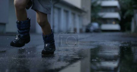 Photo for Child running into water puddle in super slow-motion. Close-up kid wearing rainboots splashing droplets after rainy afternoon, nostalgic childhood scene - Royalty Free Image